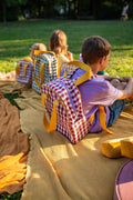 little kids sitting on blanket during picnic with green checkered backpack and red checkered backpack. gingham backpacks